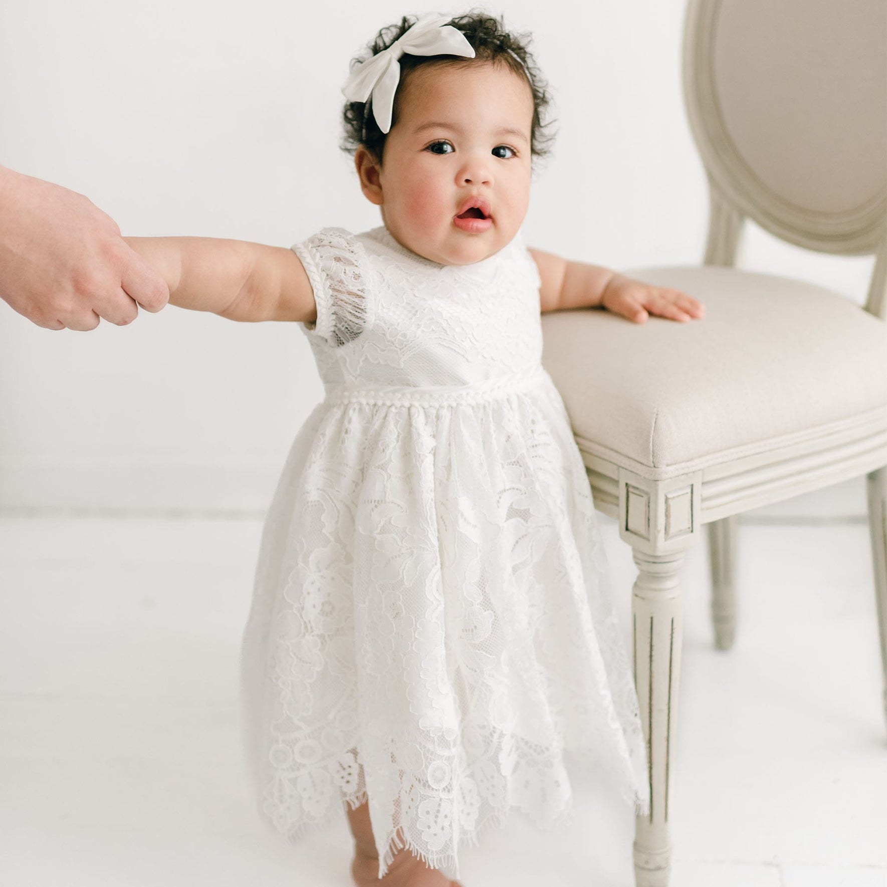 A baby wearing the Victoria Puff Sleeve Christening Dress and the Victoria Silk Bow Headband stands next to a beige chair. She is being held by the hand of someone out of the frame.