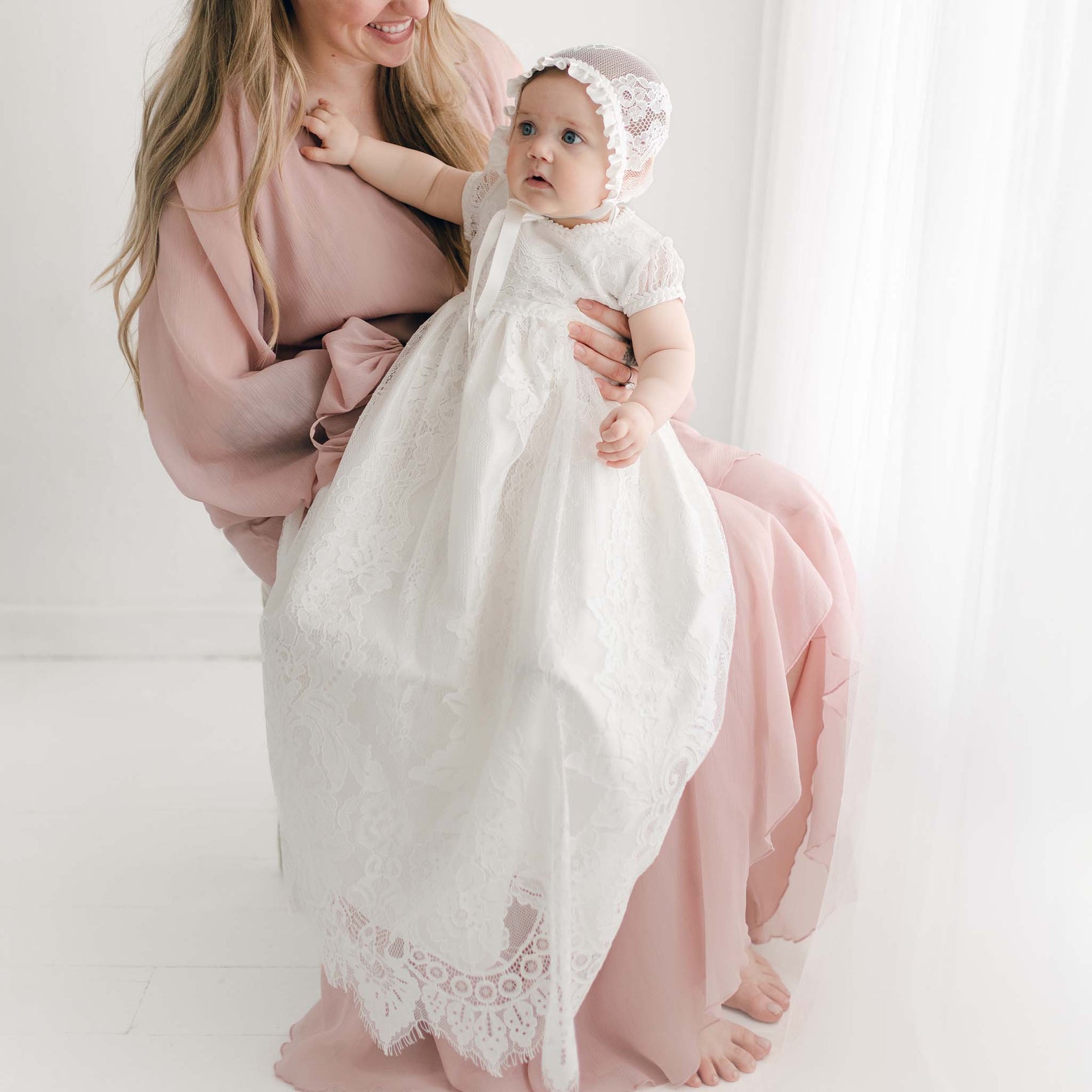 A woman in a light pink dress holds a baby wearing a Victoria Puff Sleeve Christening Gown & Bonnet. They are sitting near a window with sheer white curtains. The baby is reaching upward with one arm while the woman looks at the baby.