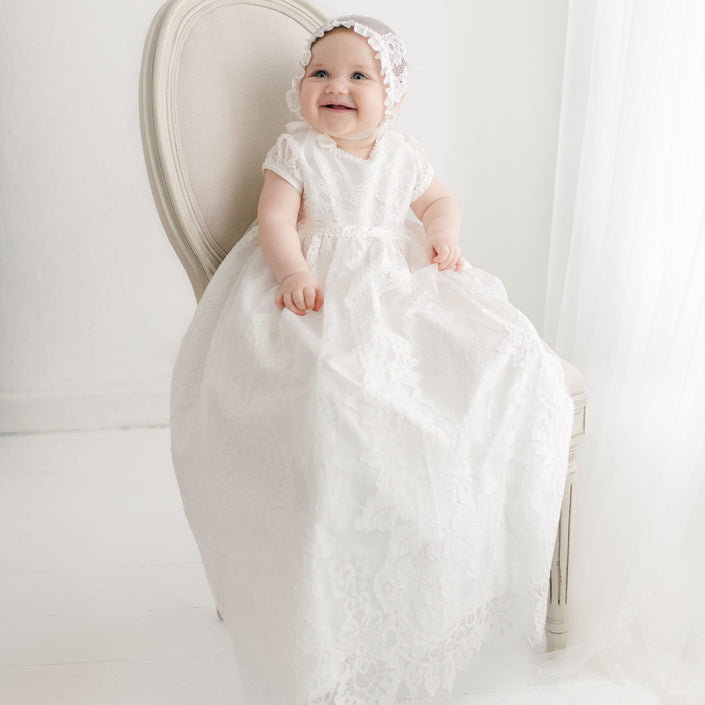 A baby is sitting on a white chair, wearing a long white Victoria Puff Sleeve Christening Gown & Bonnet with embroidered lace and a matching lace bonnet. The background is light and minimalistic with sheer white curtains. The baby is smiling and looking slightly upwards. This elegant gown, handmade in the USA, exudes timeless charm.