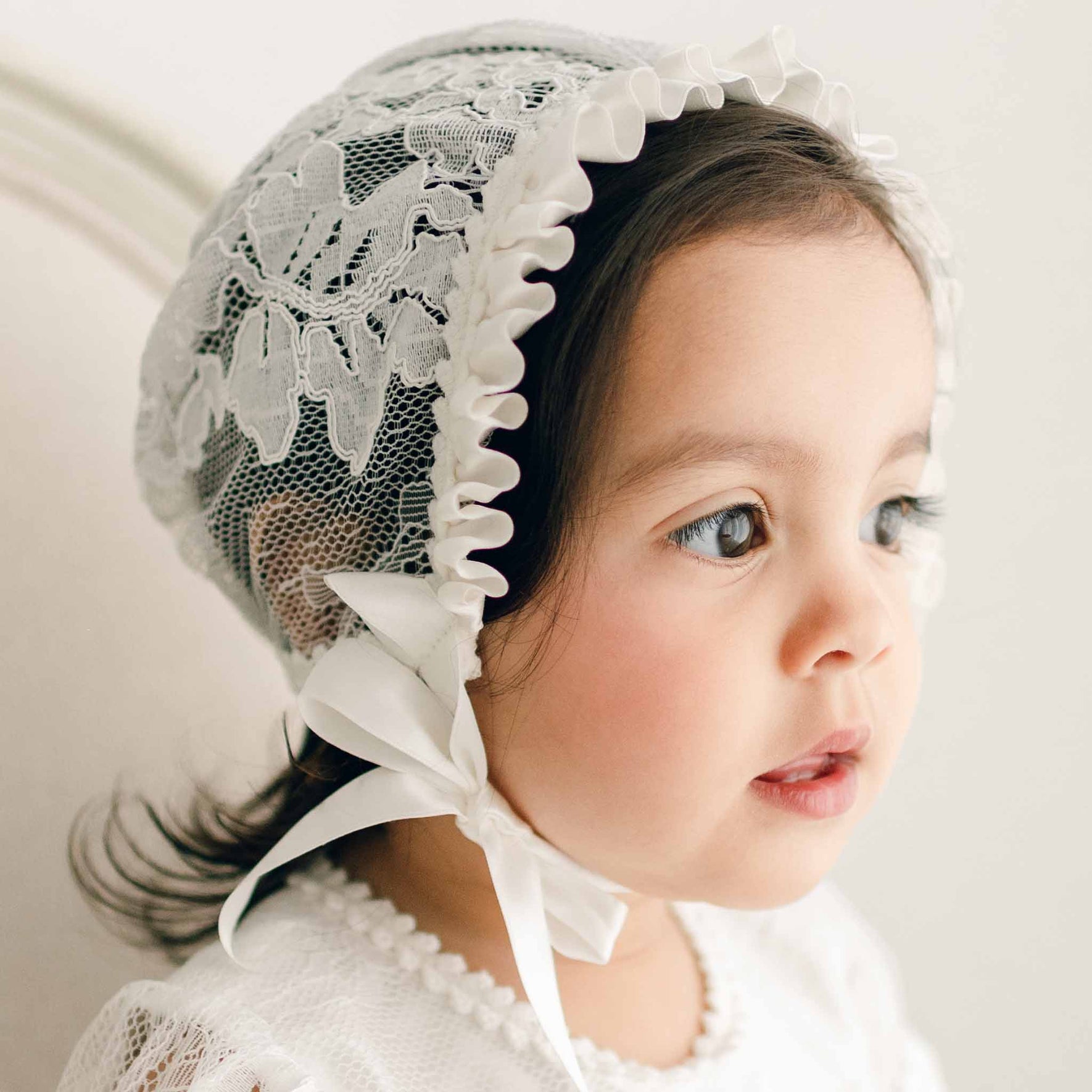 Toddler girl wearing the Victoria Lace Christening Bonnet and matching baptism gown looks to the side in profile with a thoughtful expression. This side profile highlights the intricate lacework on the bonnet.