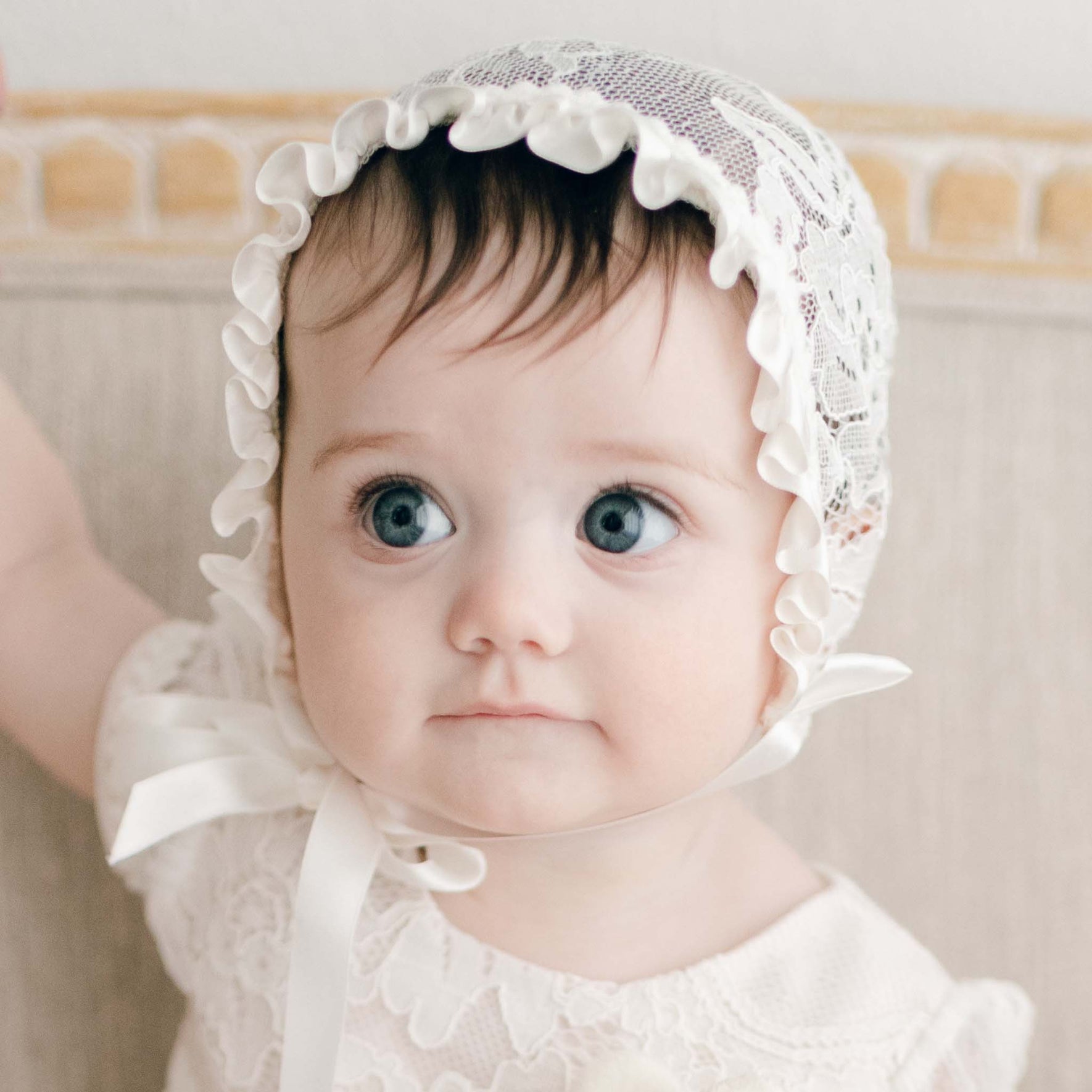 A baby girl with big blue eyes wearing the Victoria Christening Bonnet and matching baptism dress looks to the side with a slight smirk. 