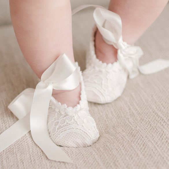 Close-up of a baby's legs and feet wearing the Victoria Lace Christening Booties. The baby is standing on a light-colored, textured fabric.