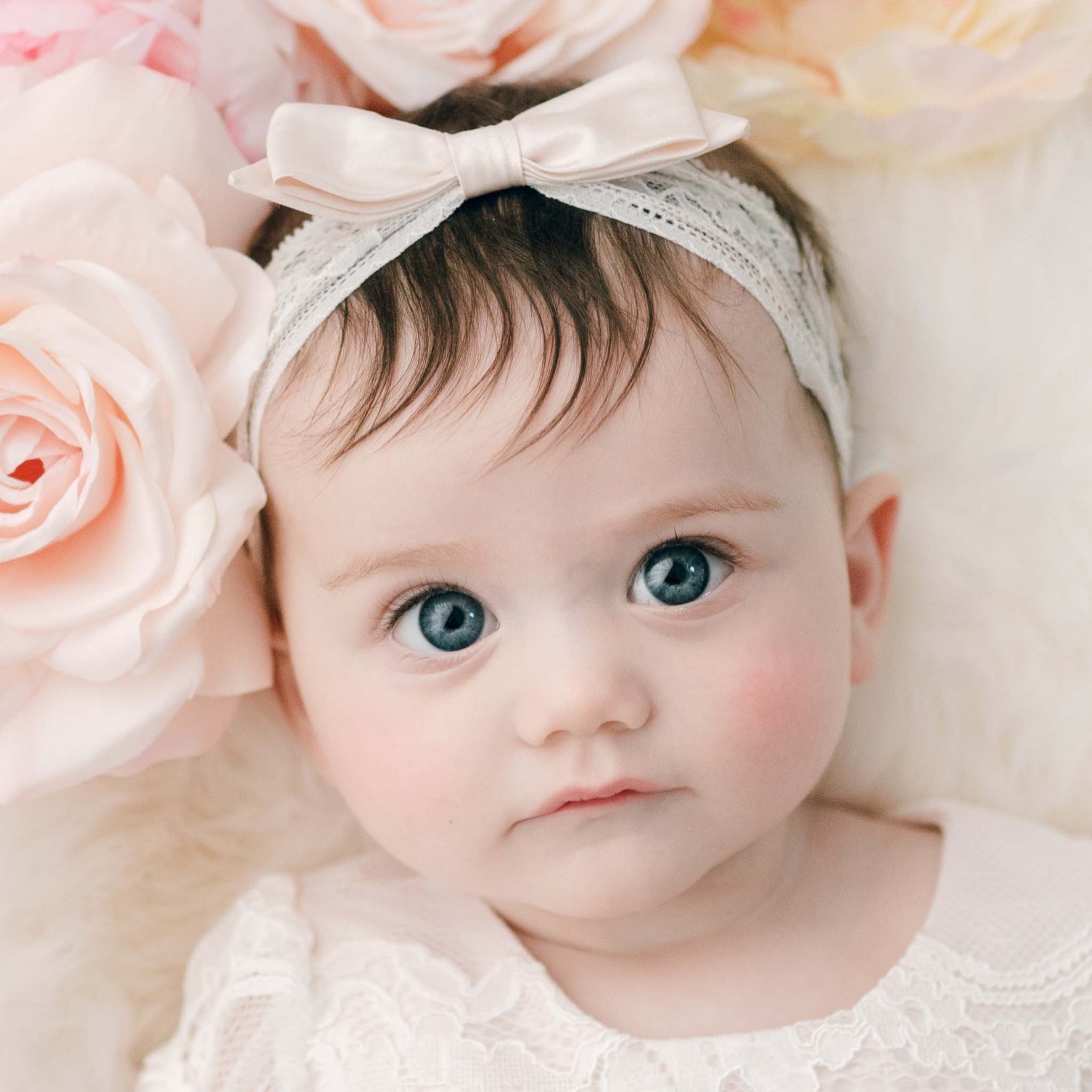 A baby with blue eyes and brown hair wears the Victoria Lace Headband, featuring a bow, while lying on a soft surface adorned with large light pink roses. Dressed in an elegant white lace outfit made from stretch floral lace, handcrafted in the USA, the little one gazes slightly to the side.