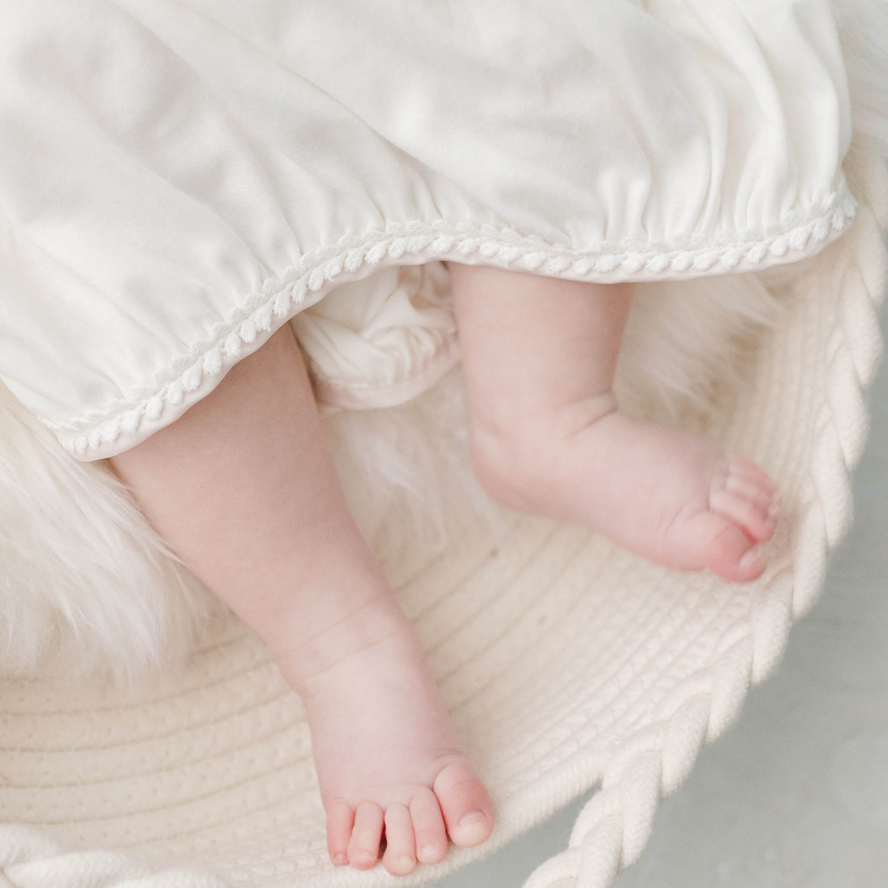 A close-up of a baby's bare feet peeking out from underneath the Victoria Layette Gown, embellished with delicate lace. The baby is resting on a soft, cream-colored surface, possibly a knit basket or cushion. The scene conveys an enchanting sense of warmth and comfort.