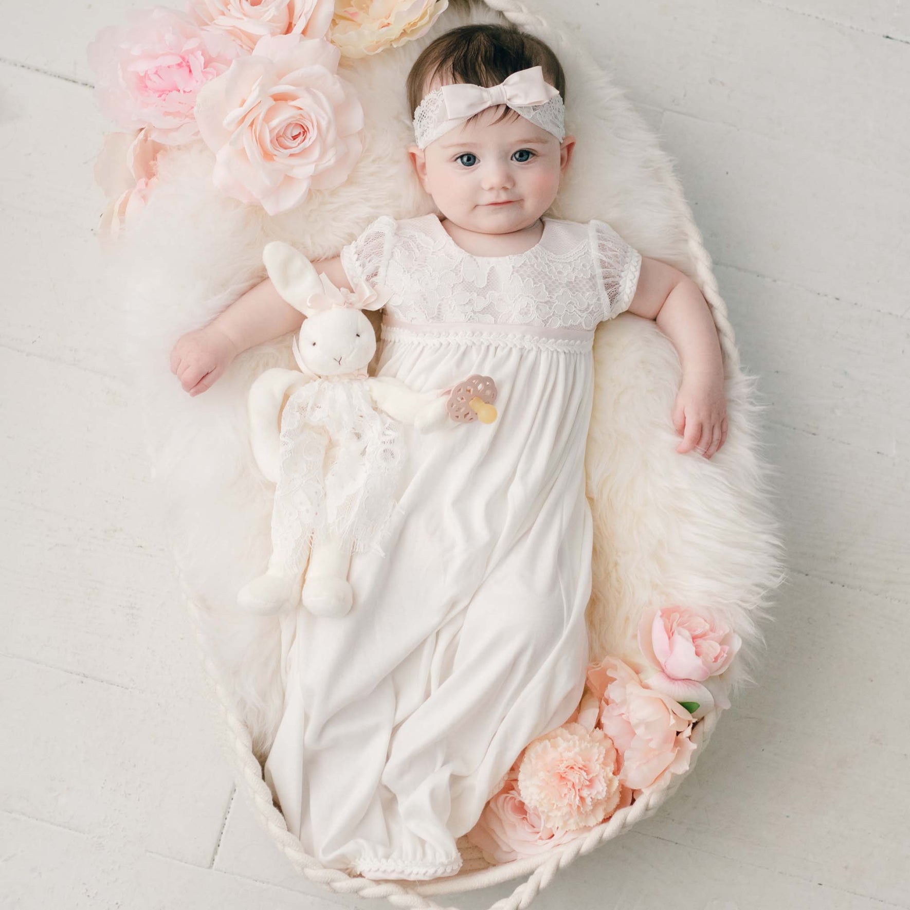 A baby dressed in a delicate Victoria Layette Gown and matching headband rests peacefully in a basket lined with a fluffy white blanket. Encircled by pink roses, the baby clutches a plush bunny toy as the serene scene unfolds on a light-colored wooden floor, capturing timeless elegance.