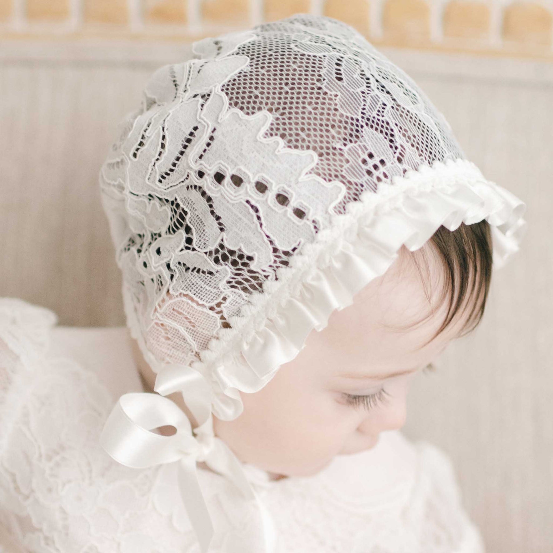 A baby is adorned with the Victoria Newborn Gift Set, which includes a white lace bonnet featuring a delicate ribbon tie and a complementary lace outfit, making it an ideal gift for a baby shower. The baby gazes downward with visible eyelashes, capturing the timeless elegance of this ensemble. The background remains soft and neutral.