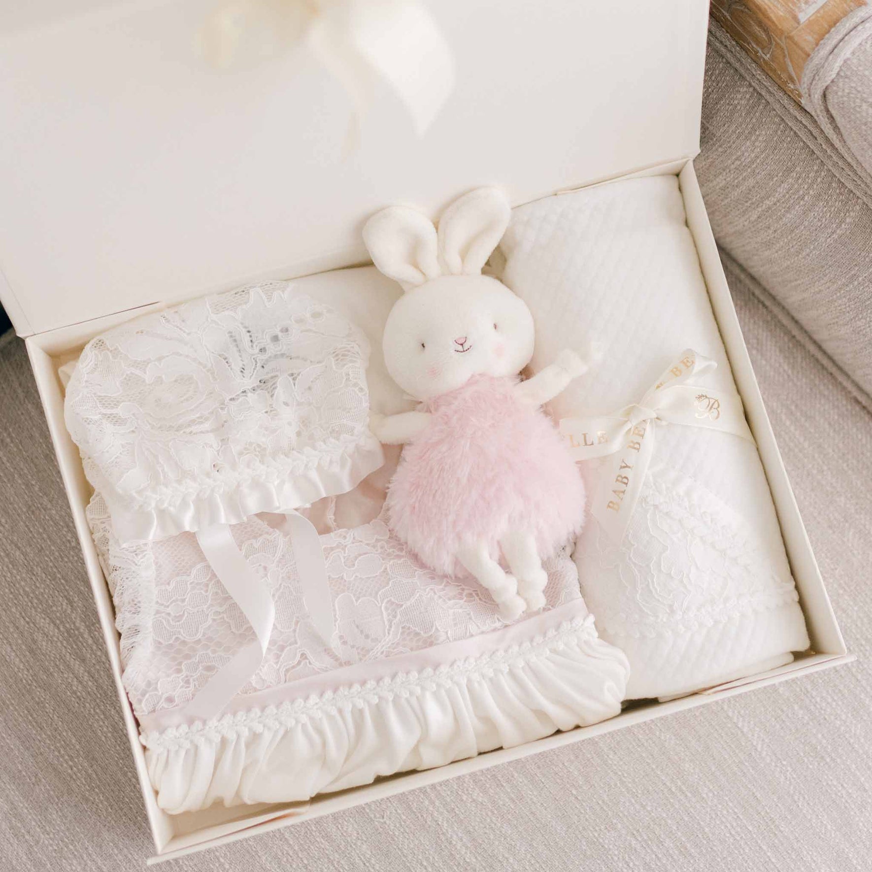 The Victoria Newborn Gift Set - Save 10% is an ideal choice for a baby shower gift, featuring a lace bonnet, a fluffy white stuffed bunny toy dressed in pink, and a textured white blanket. Each item is beautifully arranged with the blanket wrapped in a ribbon adorned with the words "Baby Baby.