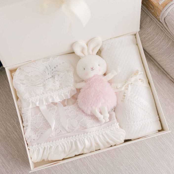 The Victoria Newborn Gift Set - Save 10% is an ideal choice for a baby shower gift, featuring a lace bonnet, a fluffy white stuffed bunny toy dressed in pink, and a textured white blanket. Each item is beautifully arranged with the blanket wrapped in a ribbon adorned with the words "Baby Baby.