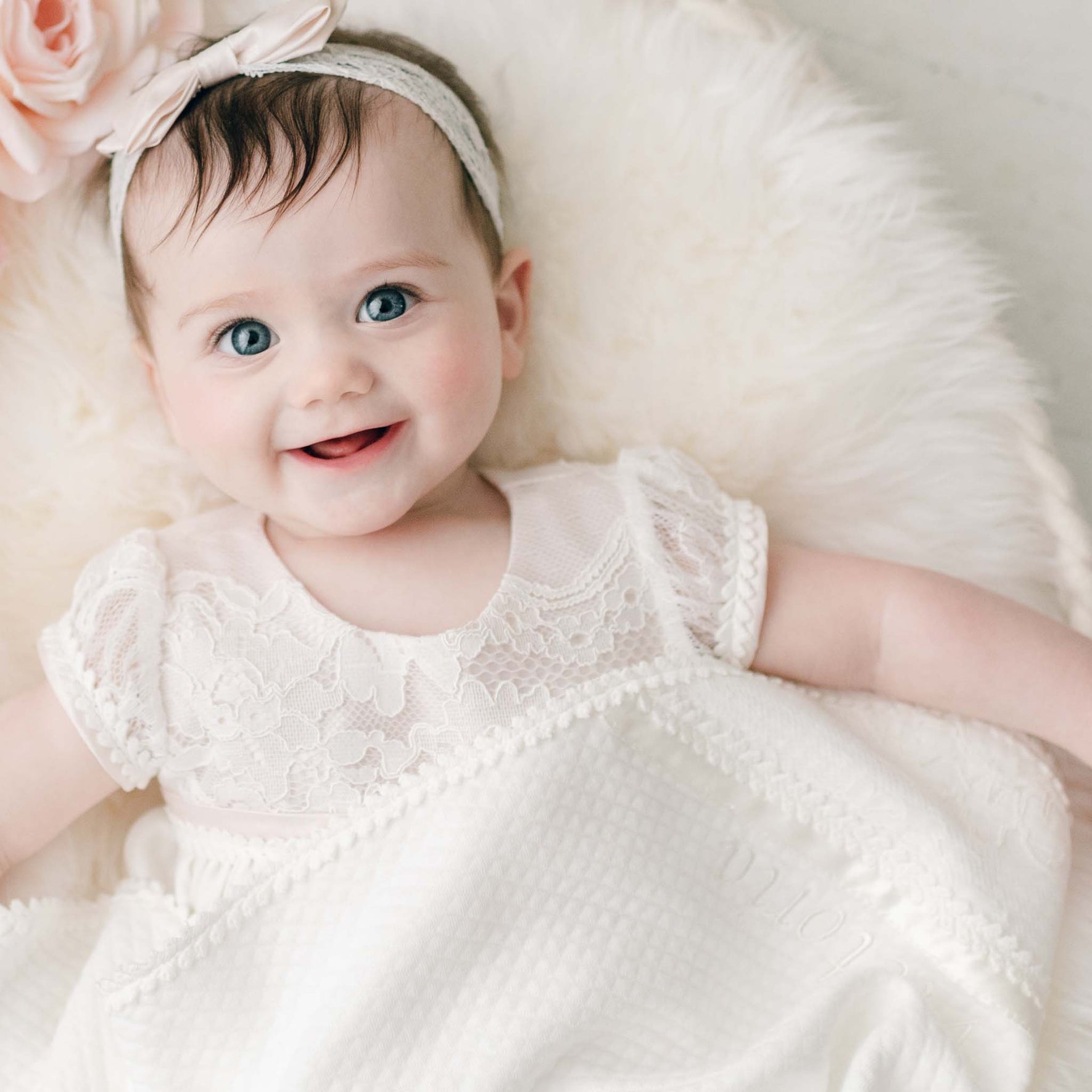 A baby with blue eyes and dark hair, dressed in an ivory embroidered lace dress and a headband with a bow, lies on the soft Victoria Personalized Blanket, smiling at the camera.