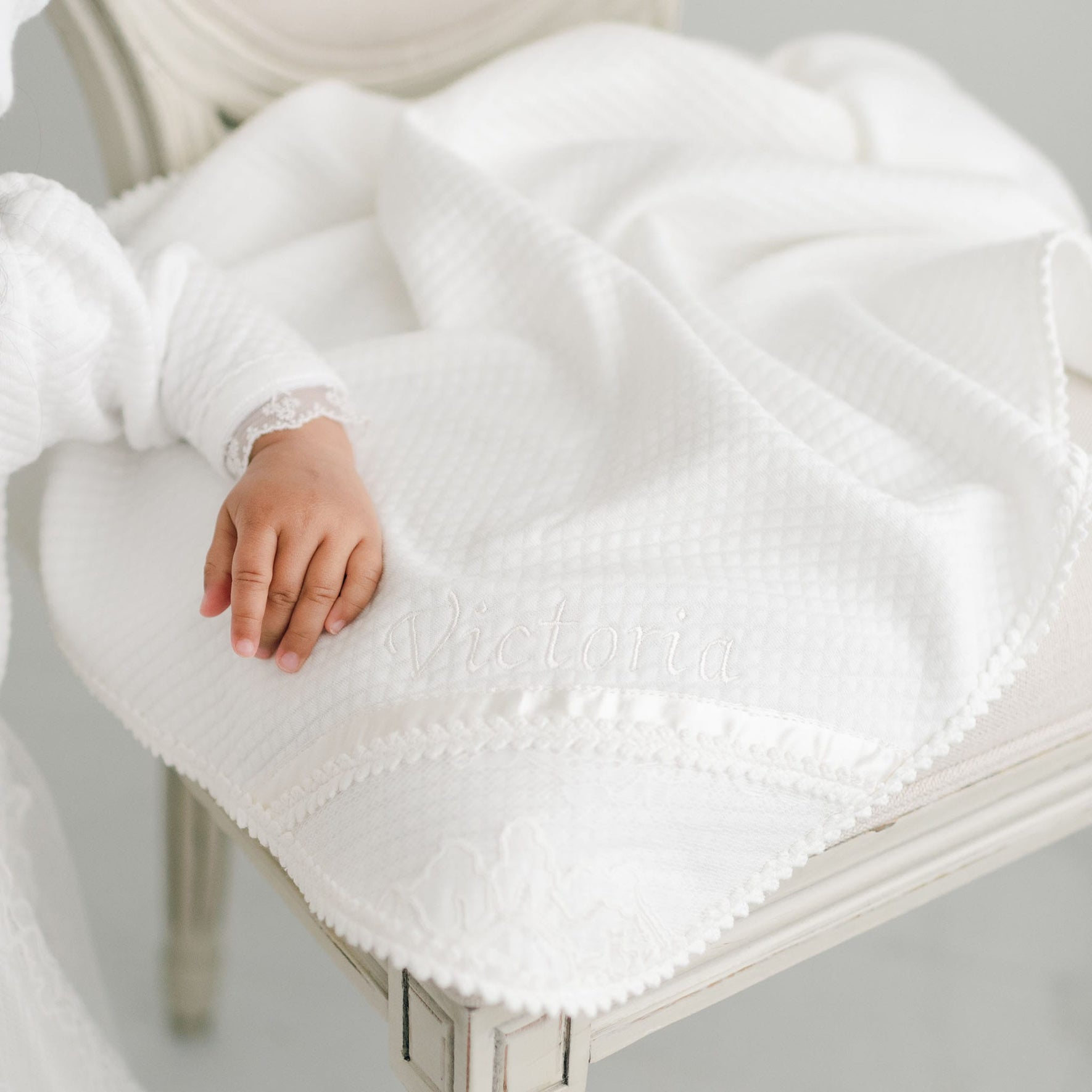 A baby's hand gently lies on a soft, white Victoria Personalized Blanket draped over a chair, crafting an ideal setting for a special gift. The exquisite christening blanket is beautifully embroidered with the name "Victoria," while the baby's sleeve showcases delicate lace trim.