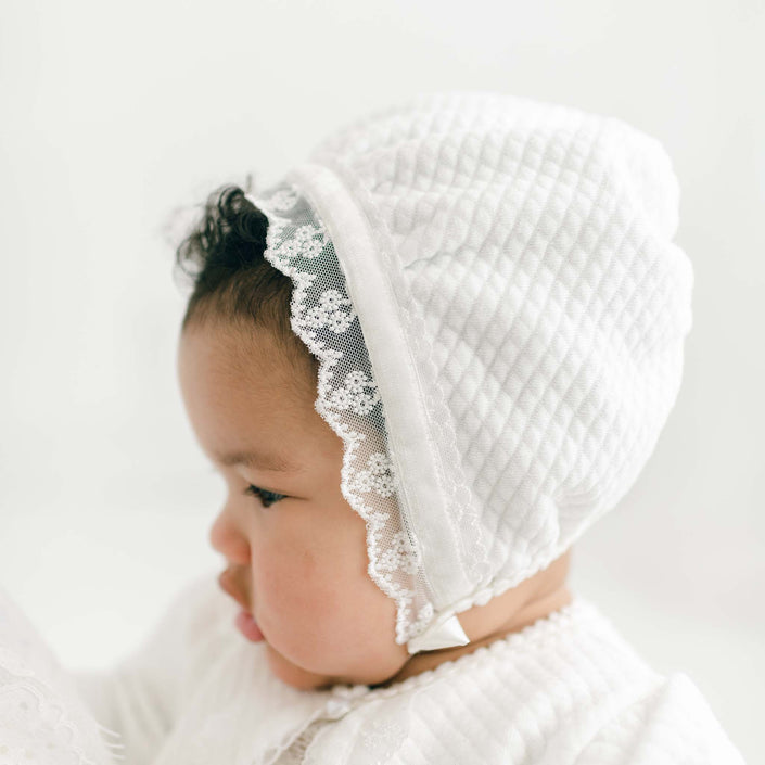 A baby wearing the Victoria Quilted Cotton Bonnet adorned with delicate ivory floral lace is seen in profile against a plain light background. The baby is also wearing the Victoria Quilted Cotton Sweater.