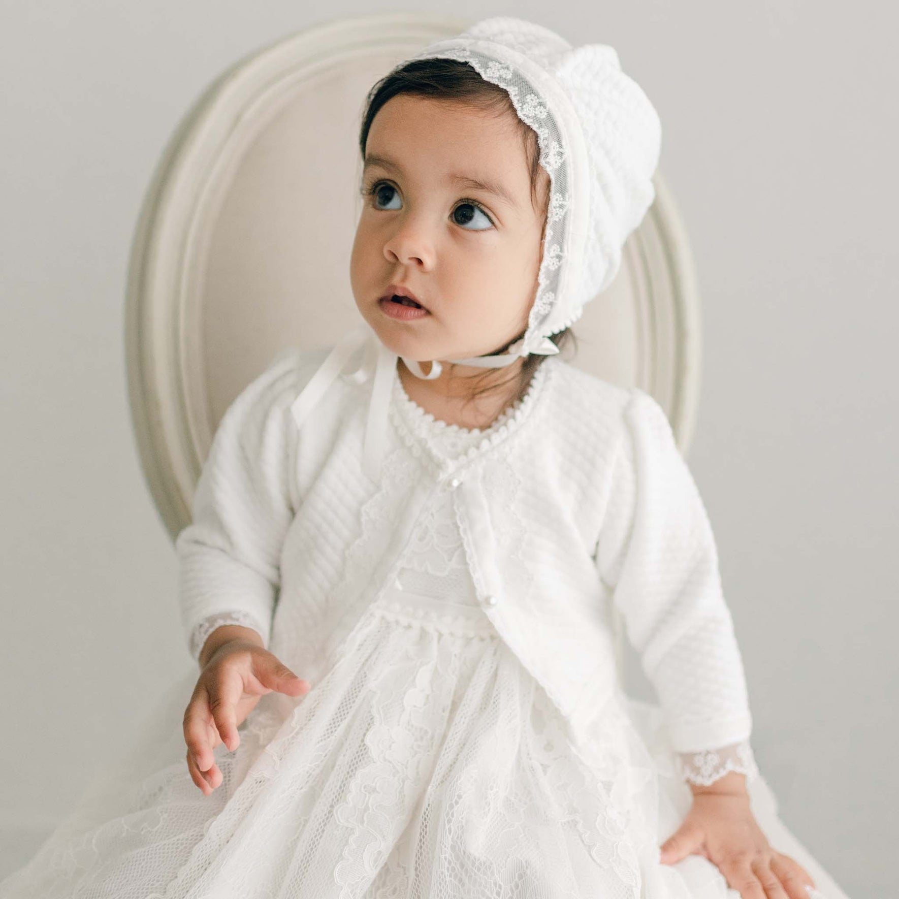 A toddler sits on a beige cushioned chair, dressed in a Victoria Quilted Cotton Bonnet and matching dress, looking slightly upward. The child has dark hair and an attentive expression. The background is light gray.