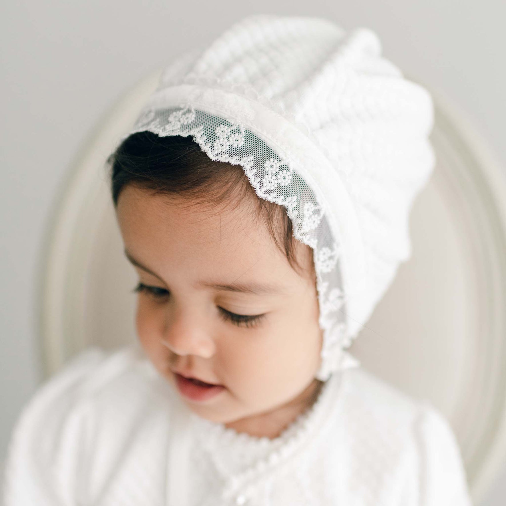A baby wearing a Victoria Quilted Cotton Bonnet with ivory floral lace trim looks downward. The child is dressed in a quilted cotton outfit and is shown in a close-up shot against a neutral background.