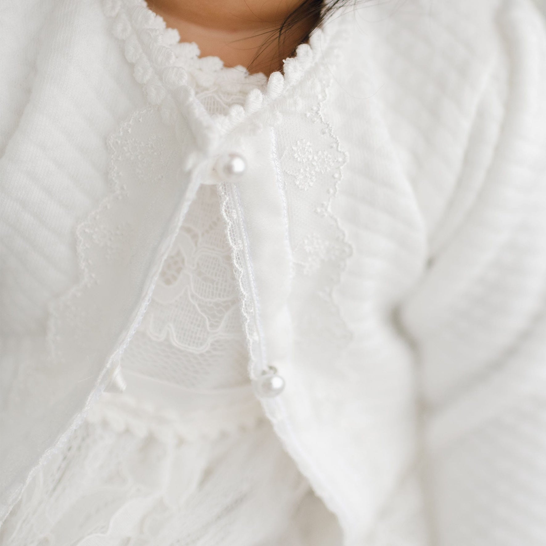 Close-up of a baby wearing a soft white Victoria Quilted Cotton Sweater over a lace dress with delicate flower embroidery. The handmade Victoria Quilted Cotton Sweater is buttoned with two small pearl style buttons. The baby’s chin and part of their face are slightly visible.