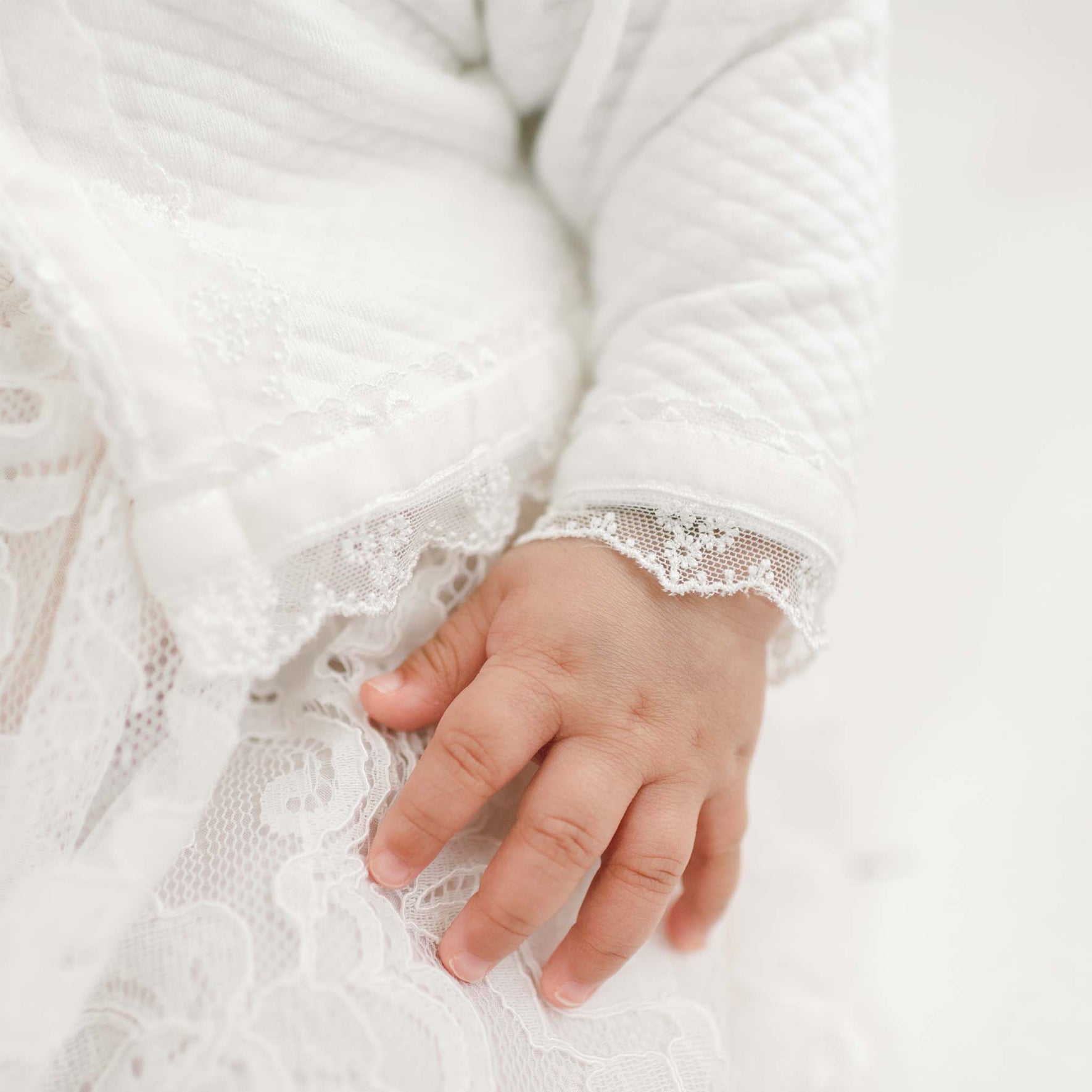Close-up of the sleeve on the Victoria Quilted Cotton Sweater. The baby's hand gently resting on the Victoria Puff Sleeve Christening Dress. The sweater features intricate lace and textured details adorned with pearl style buttons.