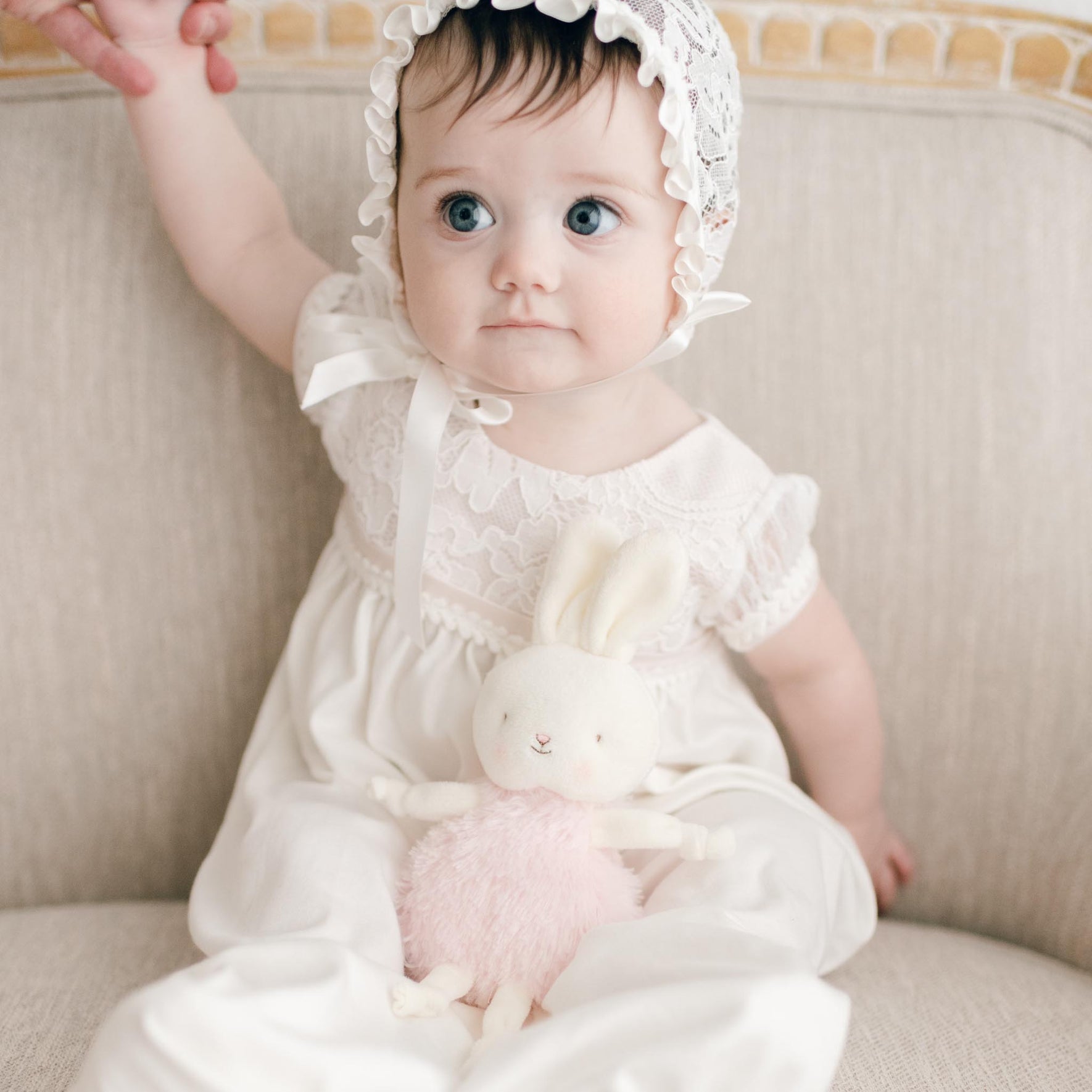A baby in a white lace bonnet and dress sits safely on a beige couch, clutching a Victoria Roly Poly Bunny with pink fur. With one arm raised and wide eyes full of wonder, the little one gazes forward.