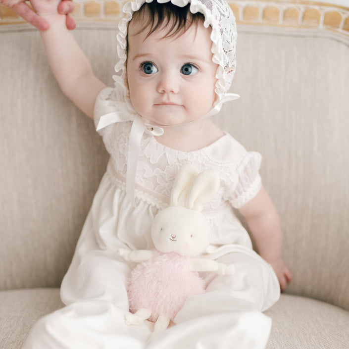 A baby in a white lace bonnet and dress sits safely on a beige couch, clutching a Victoria Roly Poly Bunny with pink fur. With one arm raised and wide eyes full of wonder, the little one gazes forward.
