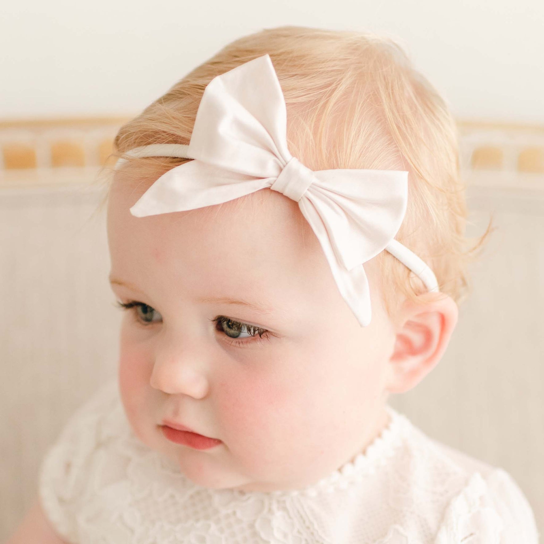 A young child with light hair is wearing a white lace outfit and a Victoria Silk Bow Headband. The child is looking slightly down and to the side, with a neutral expression. The background appears neutral and slightly blurred.