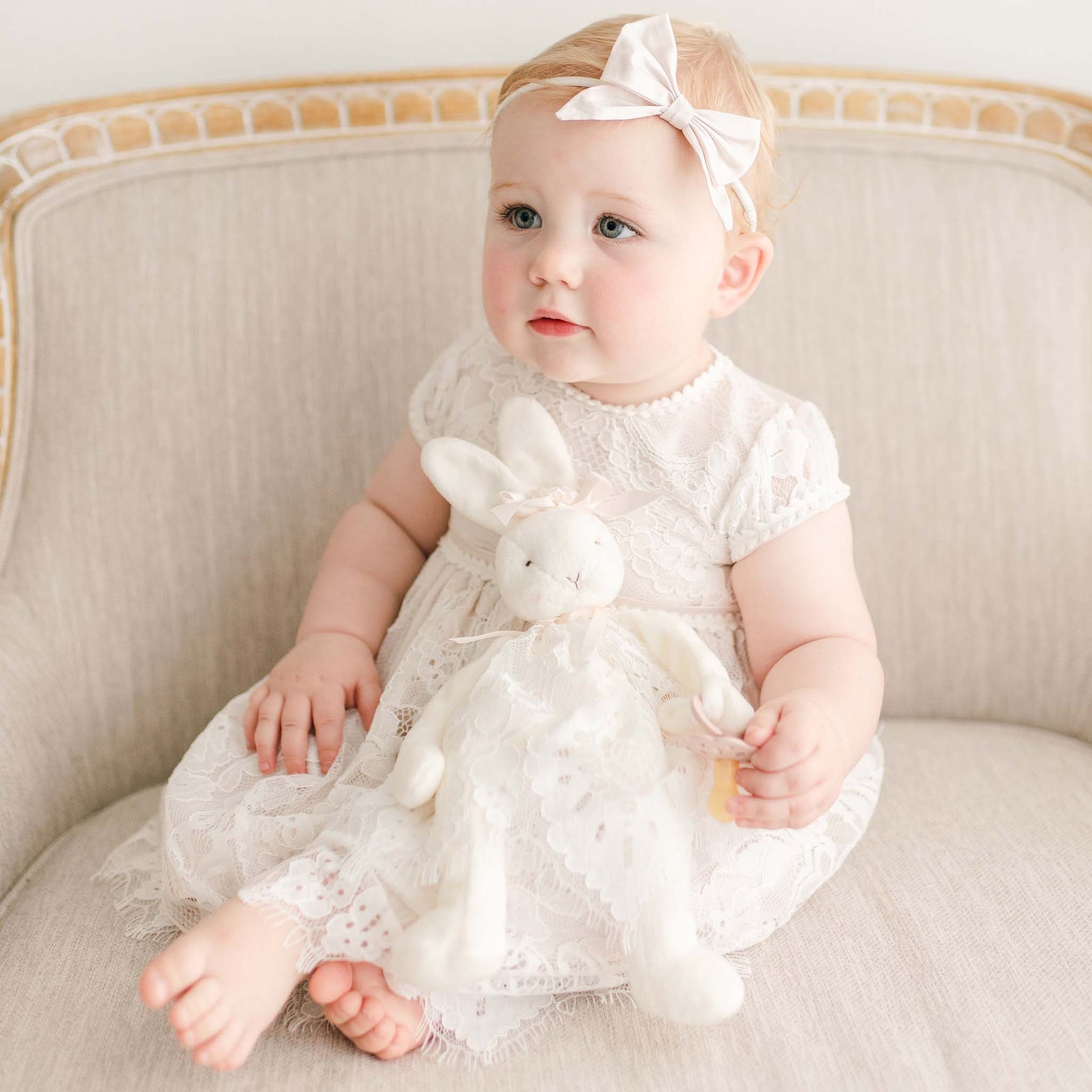 A young child with light hair sits on a beige couch, dressed in the Victoria Puff Sleeve Christening Dress with the Silk Bow Headband in their hair. They are holding the Victoria Silly Bunny Buddy. The child's expression is neutral as they look slightly off-camera.