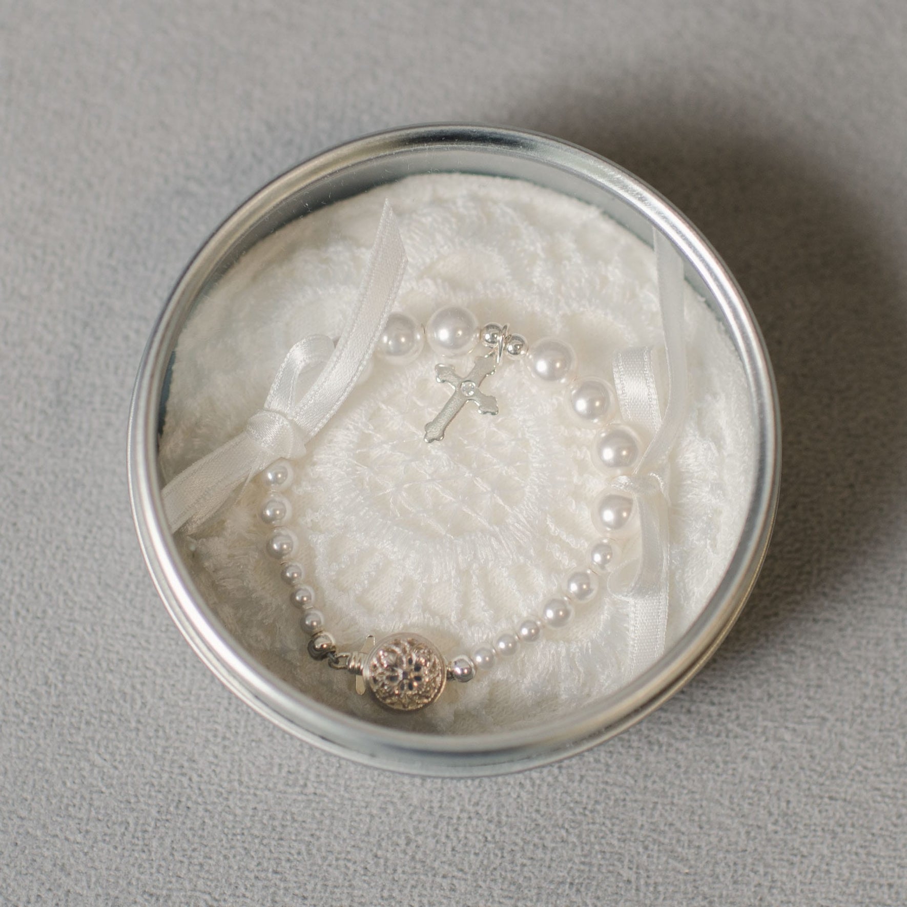 The White Luster Pearl Bracelet with Silver Cross is beautifully presented in a round silver tin featuring a delicate white lace interior and ribbon, making it an ideal Christening gift.