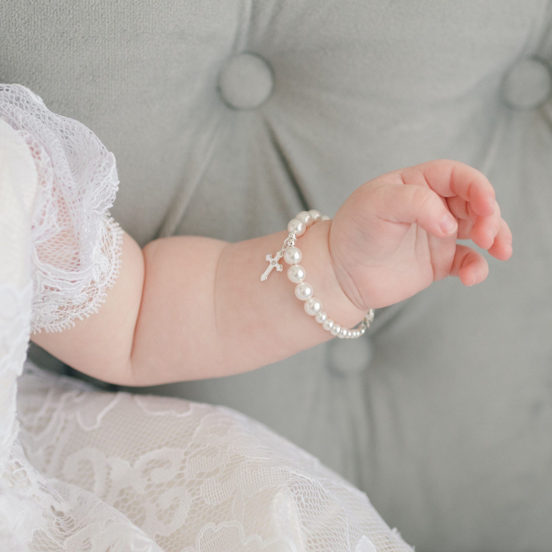 A baby's arm is visible, adorned with the White Luster Pearl Bracelet with a sterling silver cross charm. The baby, dressed in a white lace outfit, rests peacefully on a tufted gray cushion—an ideal Christening gift.