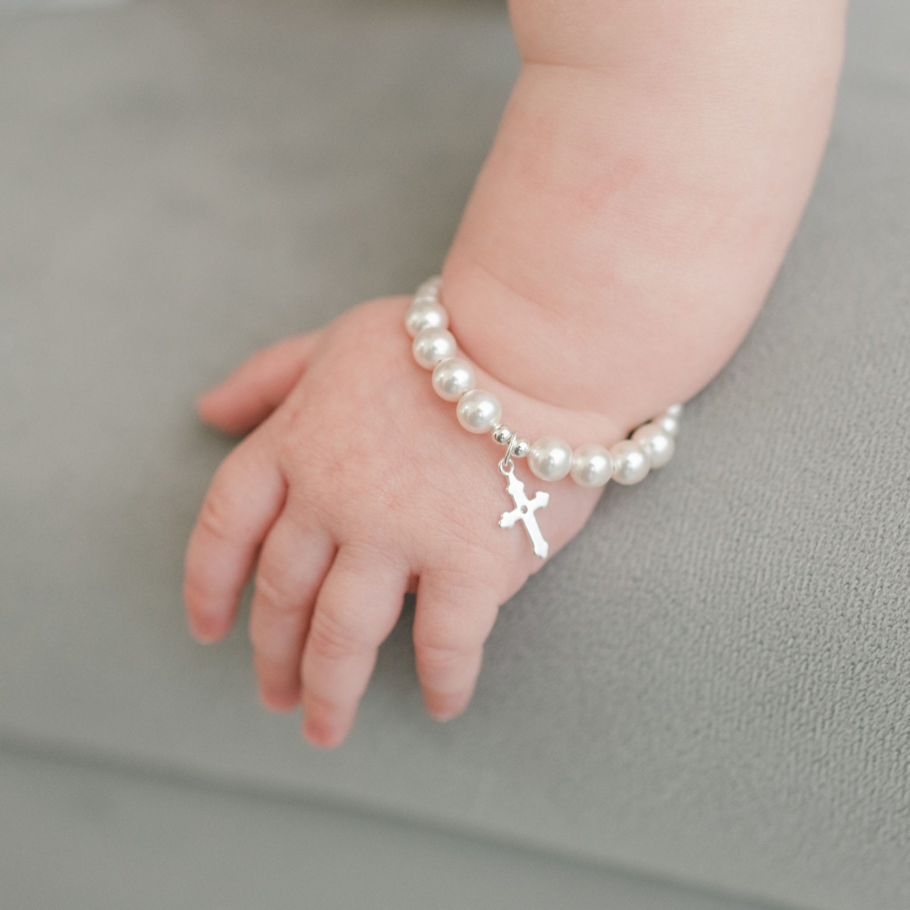 A baby's hand rests on a gray surface, adorned with the White Luster Pearl Bracelet with Silver Cross, making it a perfect Christening gift.