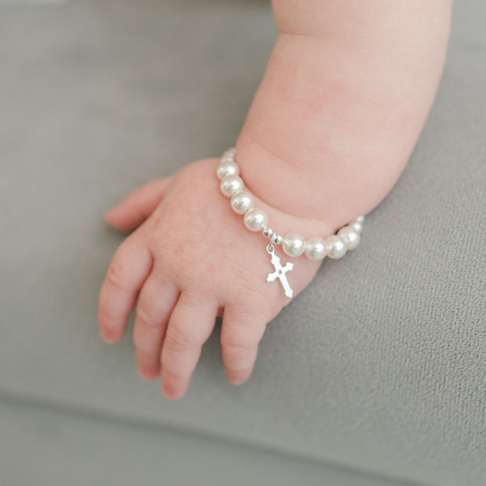 A baby's hand rests on a gray surface, adorned with the White Luster Pearl Bracelet with Silver Cross, making it a perfect Christening gift.