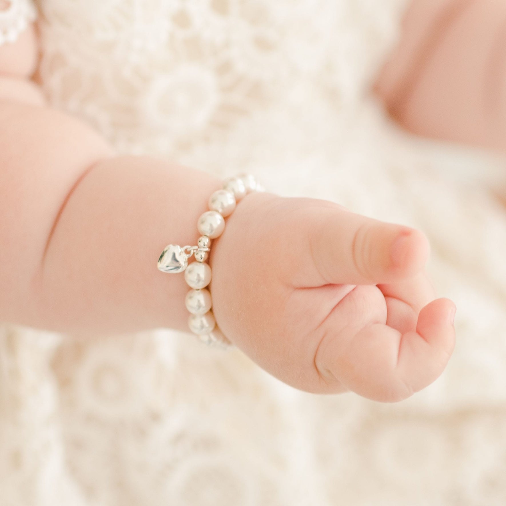 Detailed view of a baby’s hand adorned with the White Luster Pearl Bracelet featuring a silver heart charm, highlighting its elegant design.