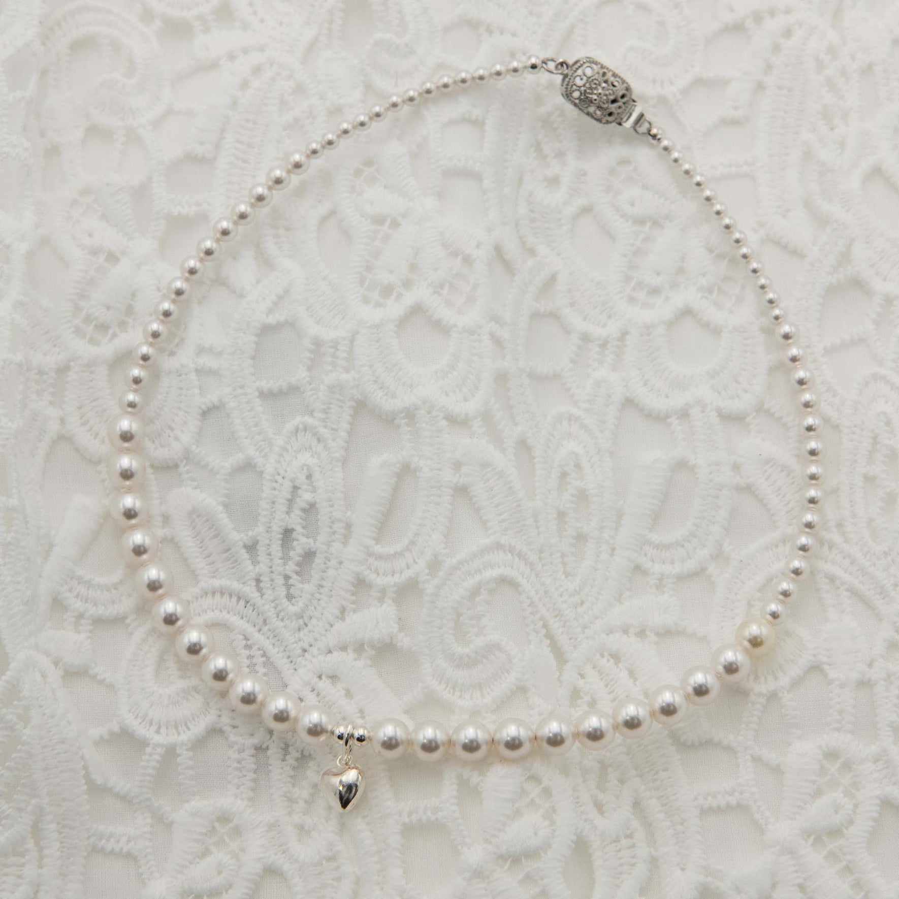 White pearl necklace featuring a small silver heart pendant, on a white lace backdrop.