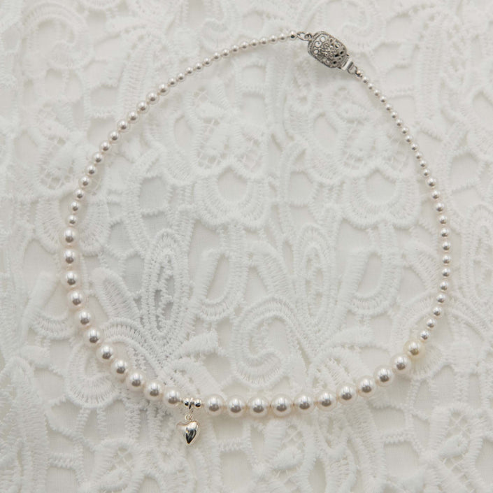 White pearl necklace featuring a small silver heart pendant, on a white lace backdrop.