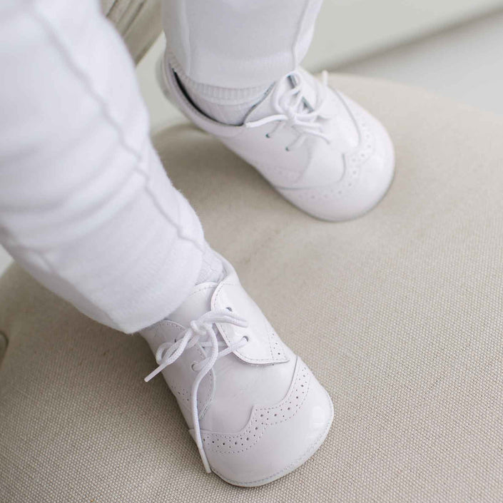 White baby shoes worn with white pants on a light-colored floor.