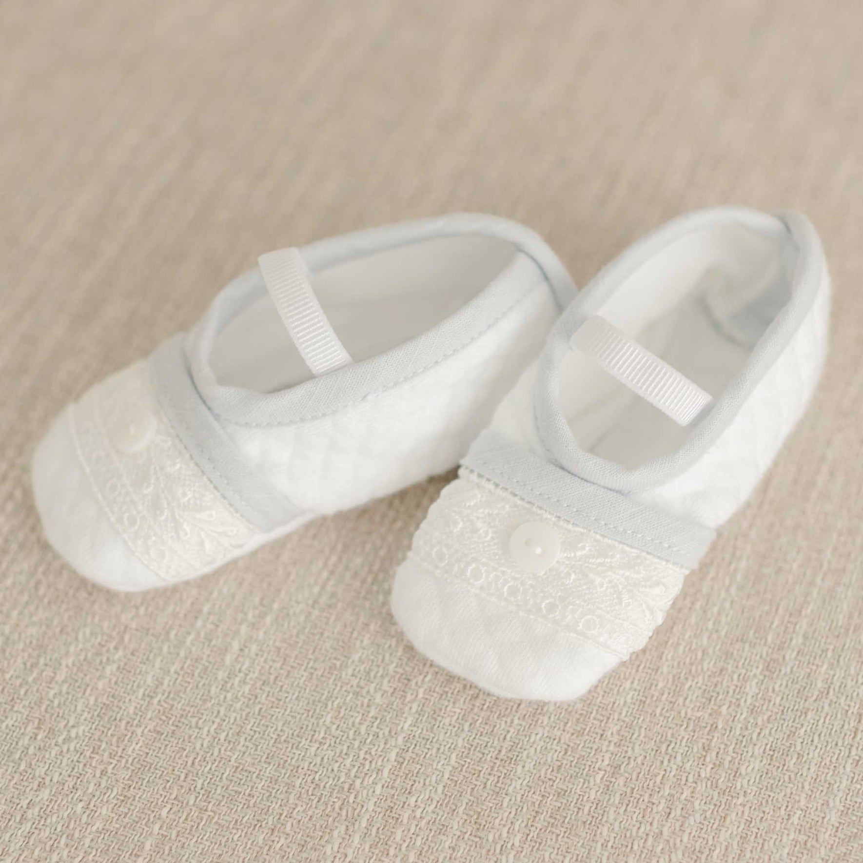 A pair of small white baby shoes featuring quilted fabric, decorative lace accents, and button details accompanies a blue knit christening sweater from the Harrison Romper Accessory Bundle - Save 15%, all placed on a beige textured surface.