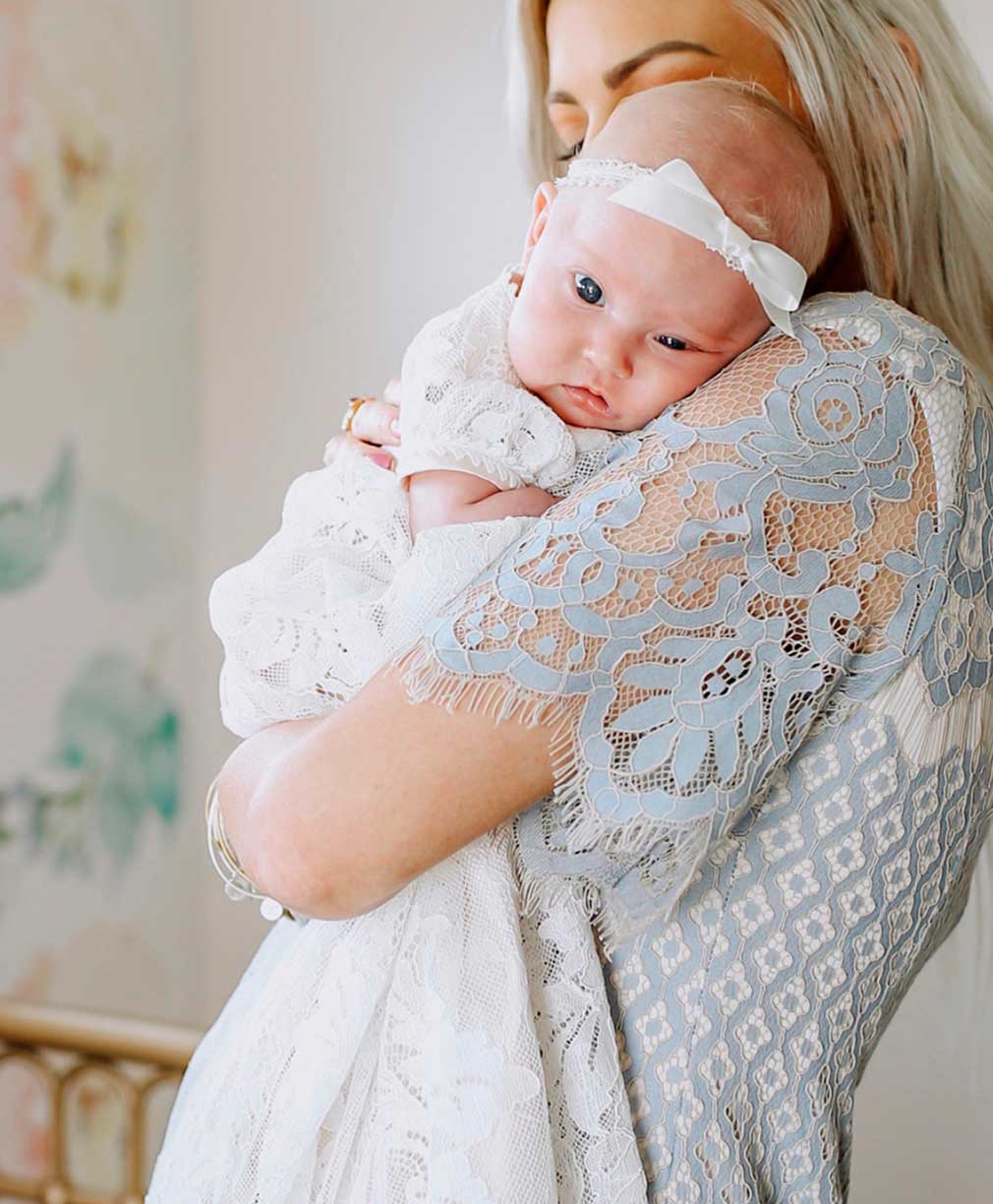 A woman is holding a baby against her shoulder. The baby is wearing a lace outfit and a white headband with a bow. The woman is dressed in a lace garment. The background is softly blurred with pastel colors.