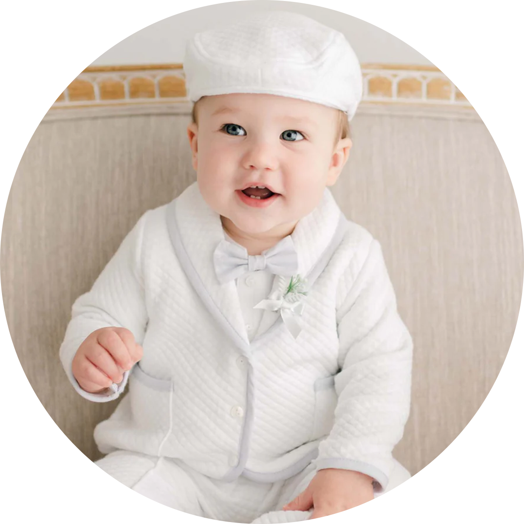 A baby wearing a white quilted outfit with a bow tie and matching cap sits on a light-colored sofa. The baby smiles while holding a white string in one hand. A small floral decoration adorns the outfit.