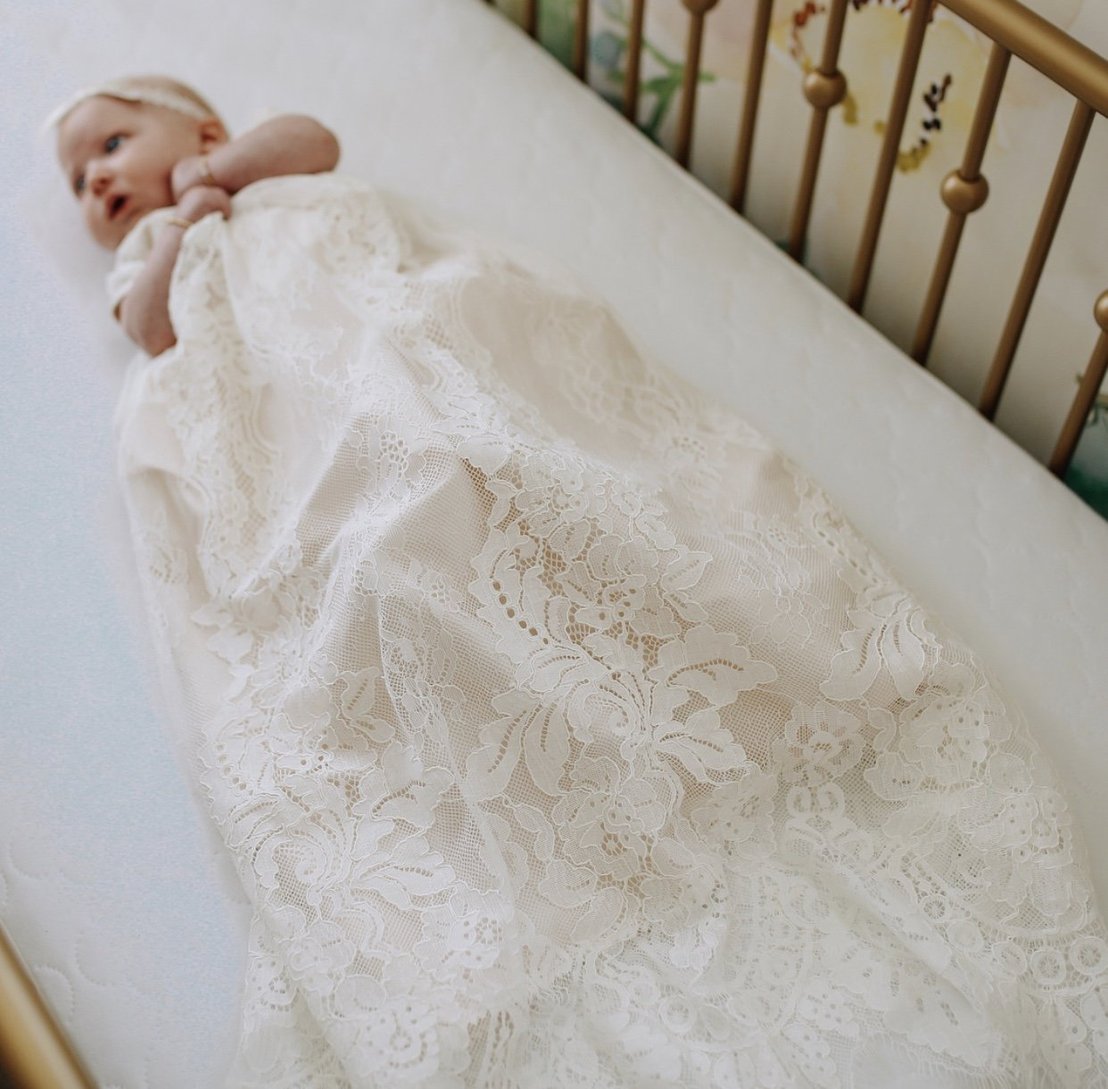 A baby wearing a long, white lace gown lies in a crib with gold-colored bars. The infant appears to be gazing upward, with hands near the face. Floral patterns decorate the background wall.