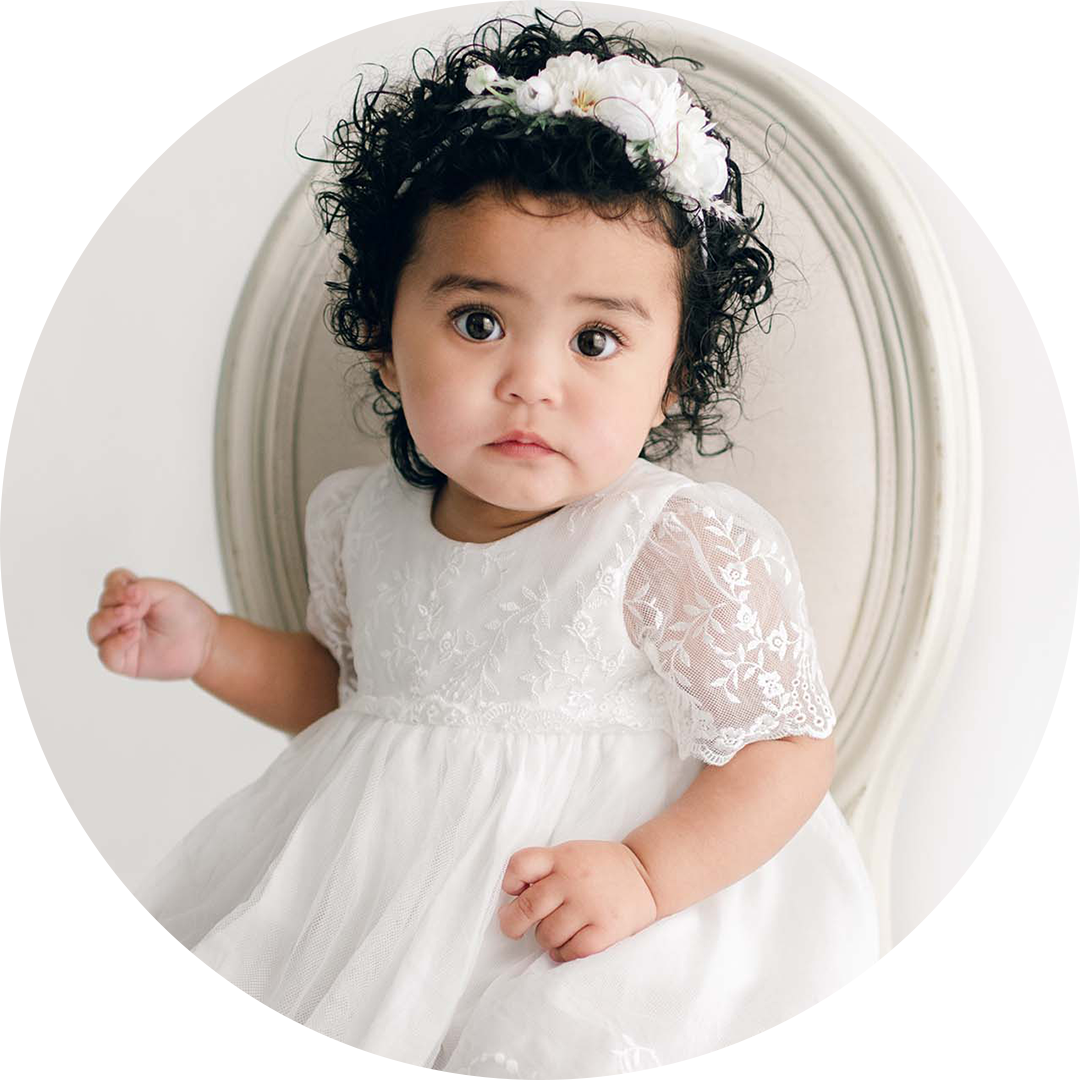 A toddler with curly hair wearing a white lace dress and a floral headband sits on a cushioned chair. She looks directly at the camera against a plain background.