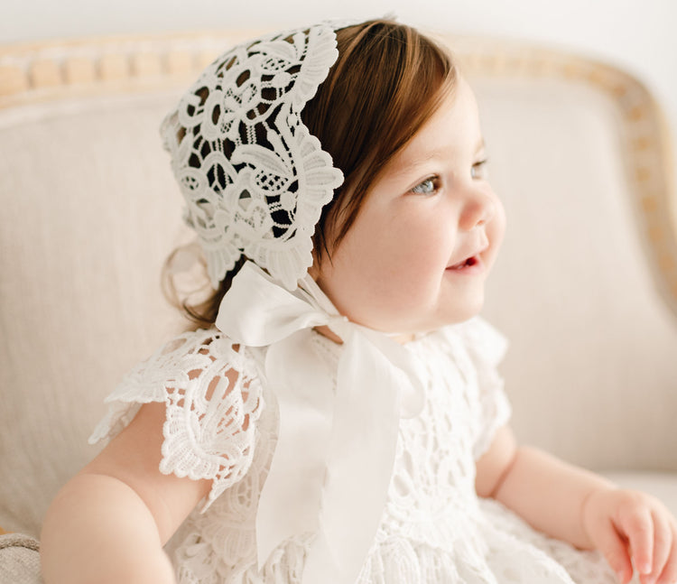 A young child wearing a white lace bonnet and dress sits on a beige couch. The child looks to the side with a slight smile, with the ribbon of the bonnet tied under the chin. The lace pattern is intricate and detailed.