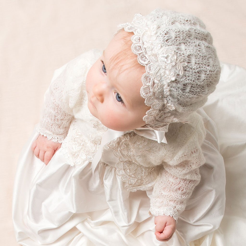 A baby is dressed in an intricate cream-colored lace gown and bonnet. The baby is sitting on a cream background, looking up with a curious expression. The outfit features detailed lace patterns and satin fabric.