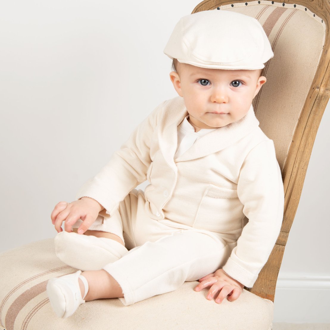 A baby sits on a wooden chair wearing a cream-colored outfit, including a cap, sweater, and pants. The background is plain white, and the baby is looking directly at the camera.