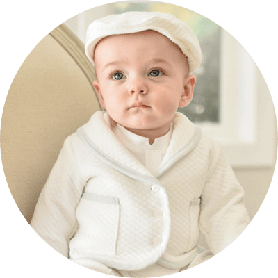 A baby wearing a white quilted outfit and matching cap sits on a beige chair. The baby has a neutral expression and looks slightly to the side. The background is softly blurred.