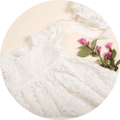 A delicate white lace dress is laid flat with intricate detailing. It is accompanied by a matching lace headband and pink flowers for decoration. The background is a soft beige color.