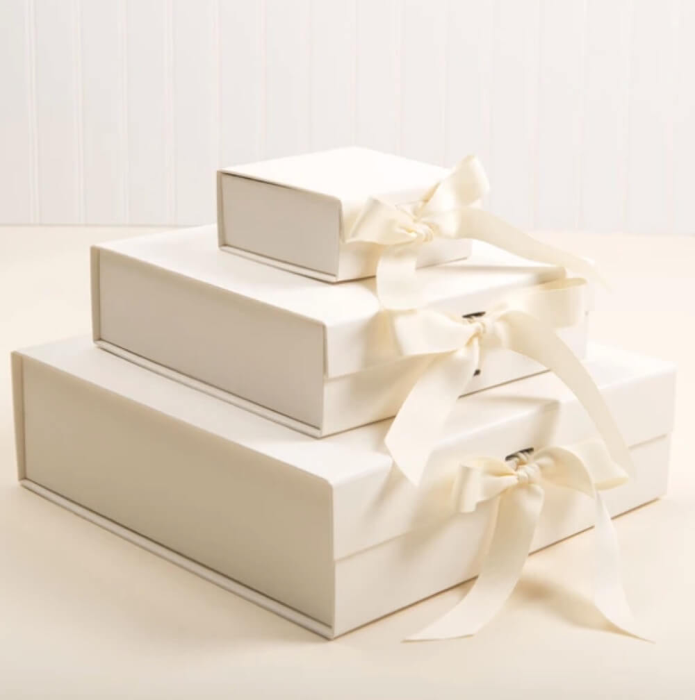 Three cream-colored gift boxes of varying sizes are stacked on top of each other. Each box is tied with a cream ribbon. The background is a light, neutral color.