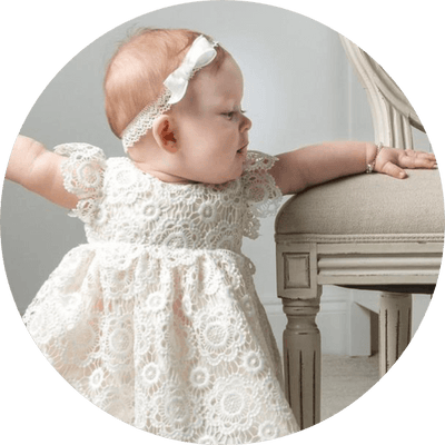 A baby wearing a white lace dress and a matching headband is standing while holding onto a beige chair. The scene is set indoors, and the baby appears to be looking towards the left.