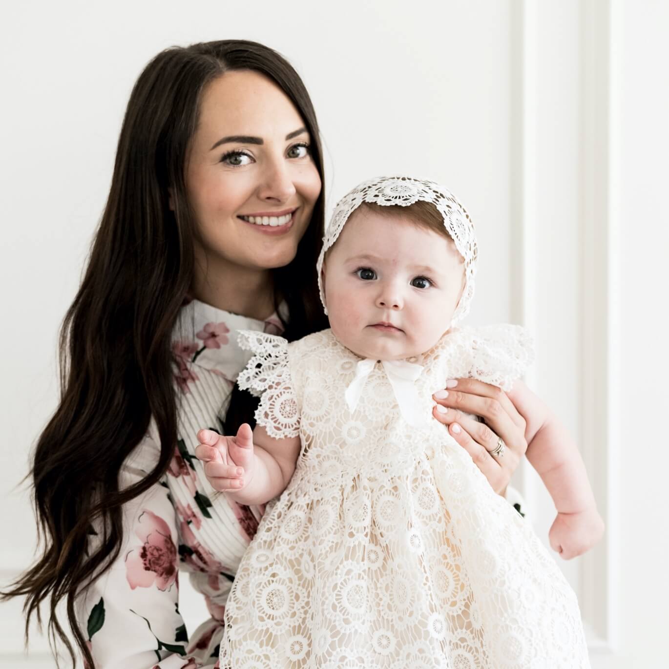 A woman with long dark hair smiles while holding a baby. The baby is wearing a white lace dress and bonnet. The woman is wearing a floral-patterned top. Both are posed against a plain light-colored background.