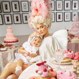 A woman in a white dress with an elaborate feathered hairstyle sits at a table, holding a child in a white outfit. The table is adorned with pink and white cakes and pastries. The background features a wall with framed pictures.