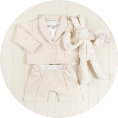 A neatly arranged baby outfit in soft cream colors is pictured, including a buttoned jacket, a pair of shorts, and a matching plush toy rabbit beside a pacifier. The background is a light-colored, vertically paneled surface.