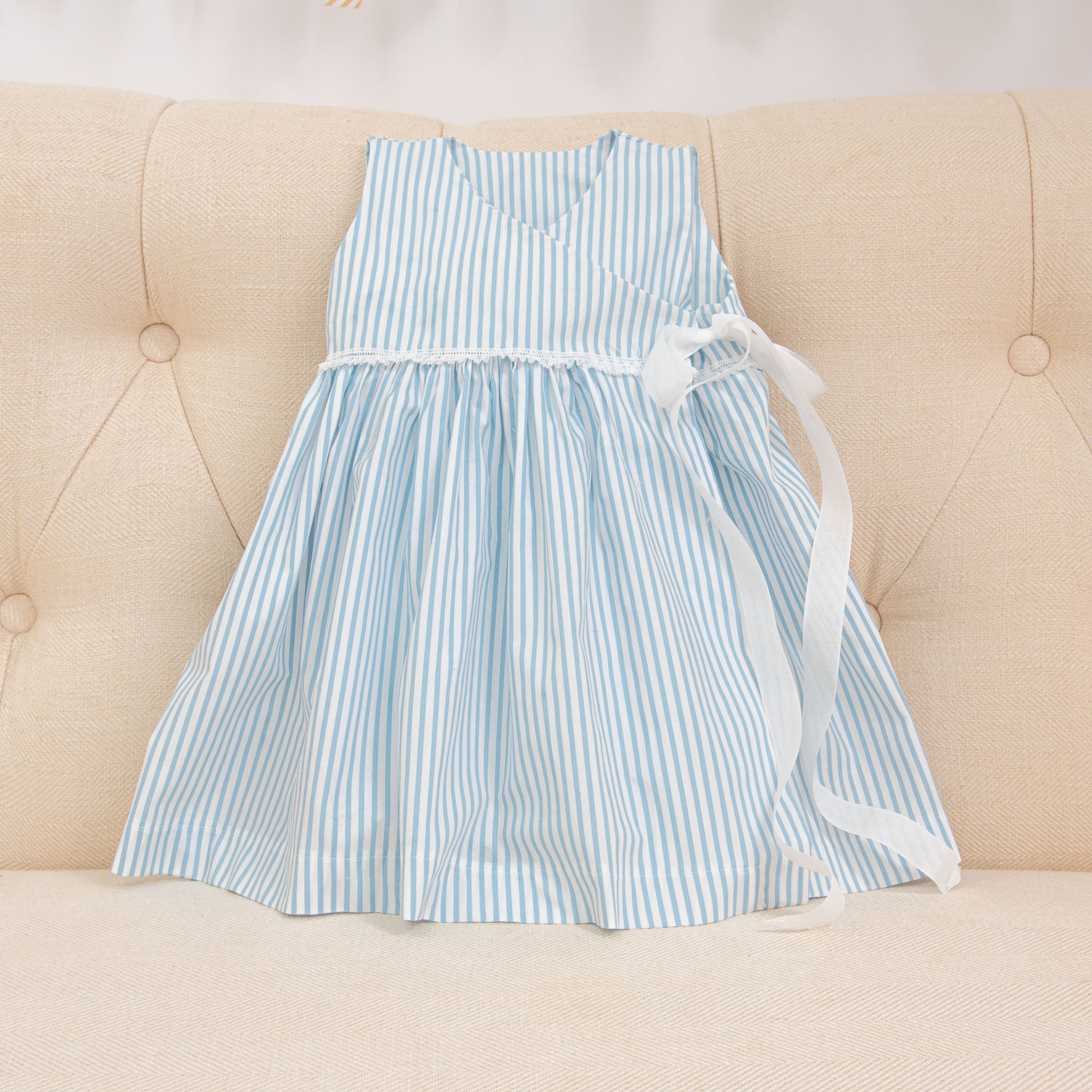 The Thea Wrap Dress, a children's cotton sleeveless outfit with light blue and white stripes, is displayed on a beige upholstered surface. It showcases a v-neckline and is adorned with a white ribbon bow at the waist, beautifully complementing the matching Thea Bow Headband.
