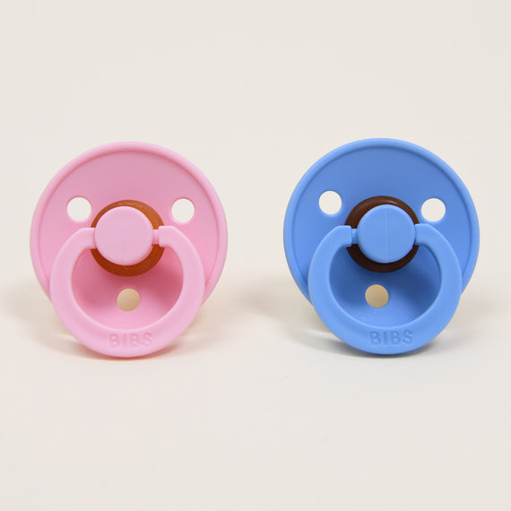 A pair of pacifiers from the Bibs Pacifier Set in Baby Pink and Sky Blue are positioned side by side against a simple white background. Made from natural rubber, each pacifier is designed with a circular shield adorned with "BIBS" text at the bottom, ideal for aiding infants in self-soothing. Crafted with attention to detail in Denmark.