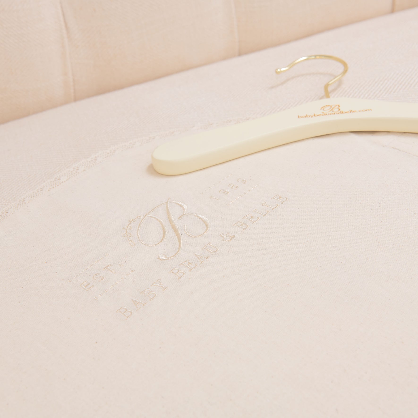 A Muslin Keepsake Garment Bag with a website URL rests on cream-colored fabric, featuring embossed text that reads "EST. 1984 BABY BEAU & BELLE," delicately complemented by lace edging. The cushioned upholstery serves as an elegant backdrop.