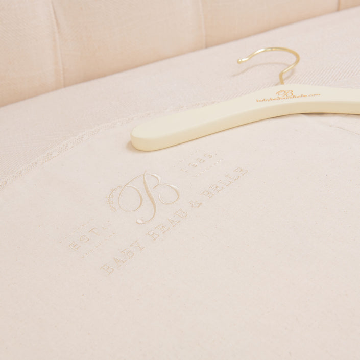 A Muslin Keepsake Garment Bag with a website URL rests on cream-colored fabric, featuring embossed text that reads "EST. 1984 BABY BEAU & BELLE," delicately complemented by lace edging. The cushioned upholstery serves as an elegant backdrop.